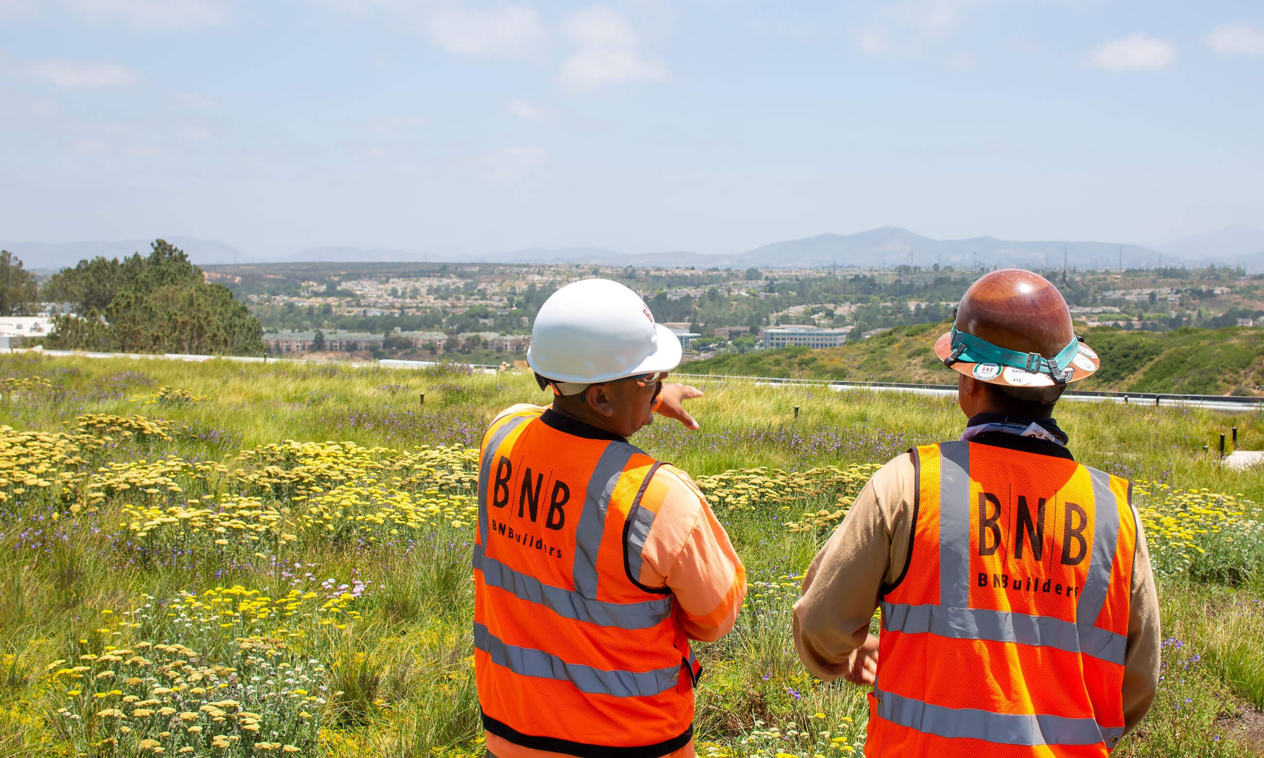 Two people with hardhats and BNBuilders orange vests looking out over a field.